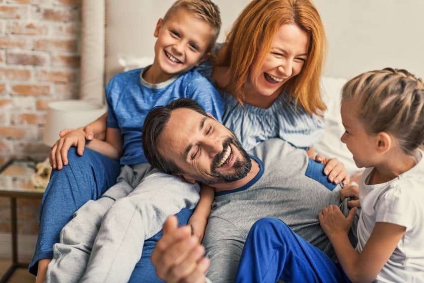 Happy family of 4 with 2 young children enjoying playtime on a couch in their living room