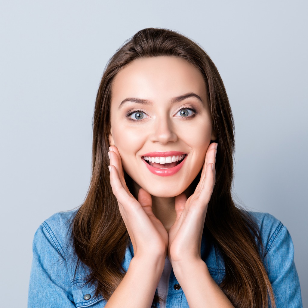 A woman in a blue shirt smiling and framing her face to show of her new smile.