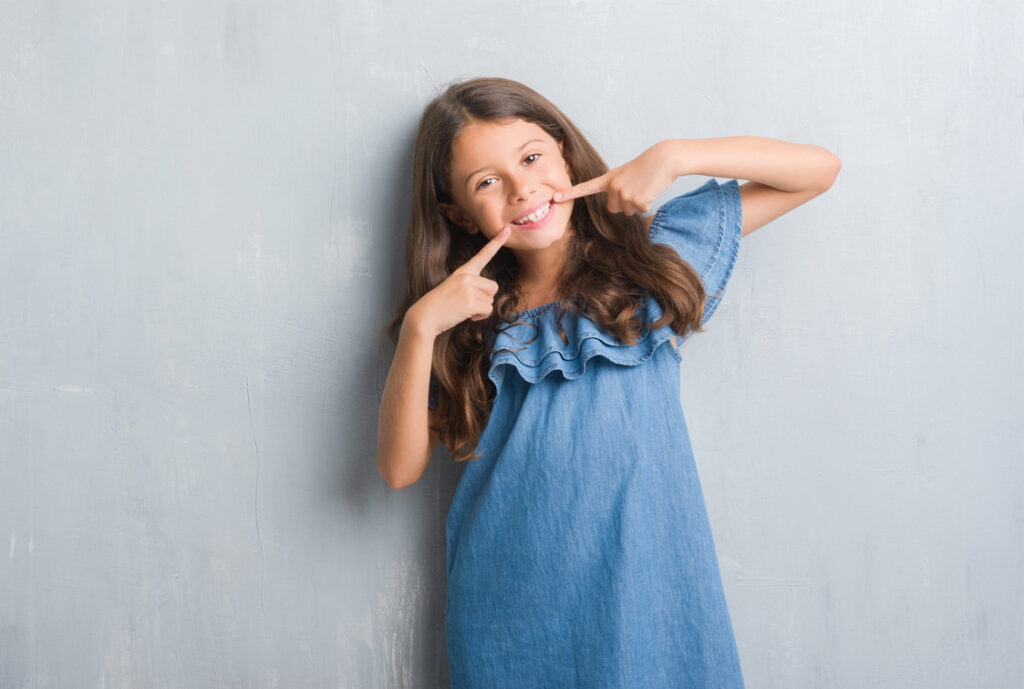 Child with brown flowing hair in a blue jean dress smiling and pointing to her teeth