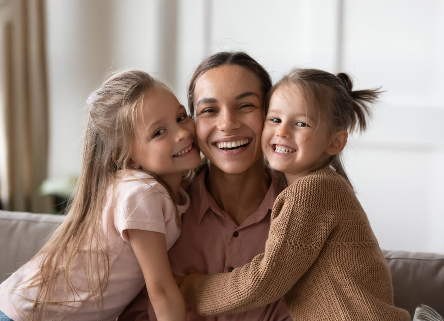 Beautiful female family young smiling single mother and cute little children daughters embracing looking at camera, funny small kids girls hugging happy mum bonding sit on sofa together, portrait